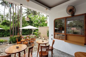 An outdoor café with wooden tables and chairs, surrounded by lush greenery and a view of the street. - Merta Luwih villa (Tabanan)