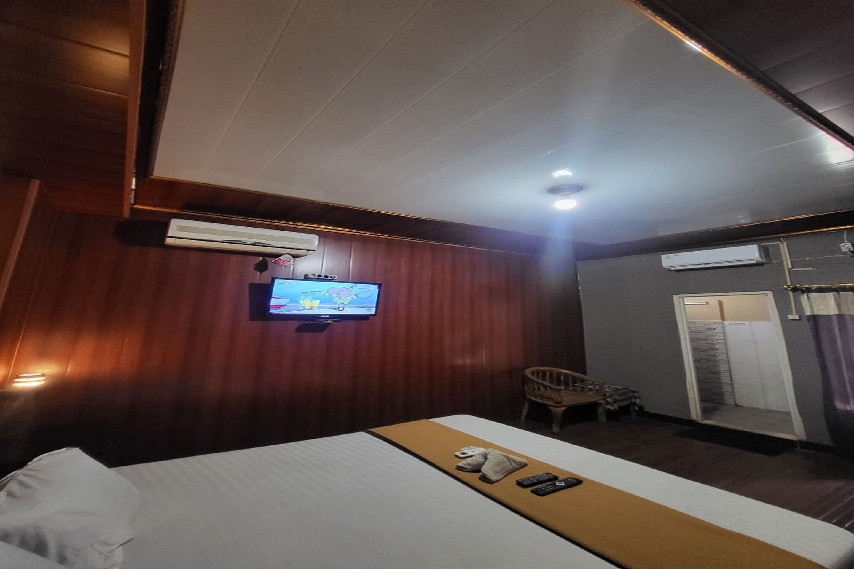 A hotel room with a bed, a television mounted on the wall, a small wooden chair, and a wardrobe.