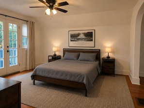 A bedroom with a bed, two bedside tables, a ceiling fan, a framed picture on the wall, and a door with curtains. - Historic Omaha Home overlooks Park. 3BR. Free Parking. 2.7 miles to Schwab Field (Omaha)