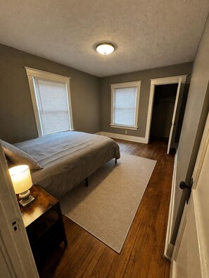 A bedroom with a bed, bedside table, lamp, two windows, a door, and a wooden floor. - Historic Omaha Home overlooks Park. 3BR. Free Parking. 2.7 miles to Schwab Field (Omaha)