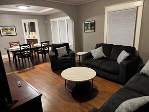 A living room with a dark-colored sofa, two armchairs, a coffee table, and a dining area with a table and chairs. - Historic Omaha Home overlooks Park. 3BR. Free Parking. 2.7 miles to Schwab Field (Omaha)