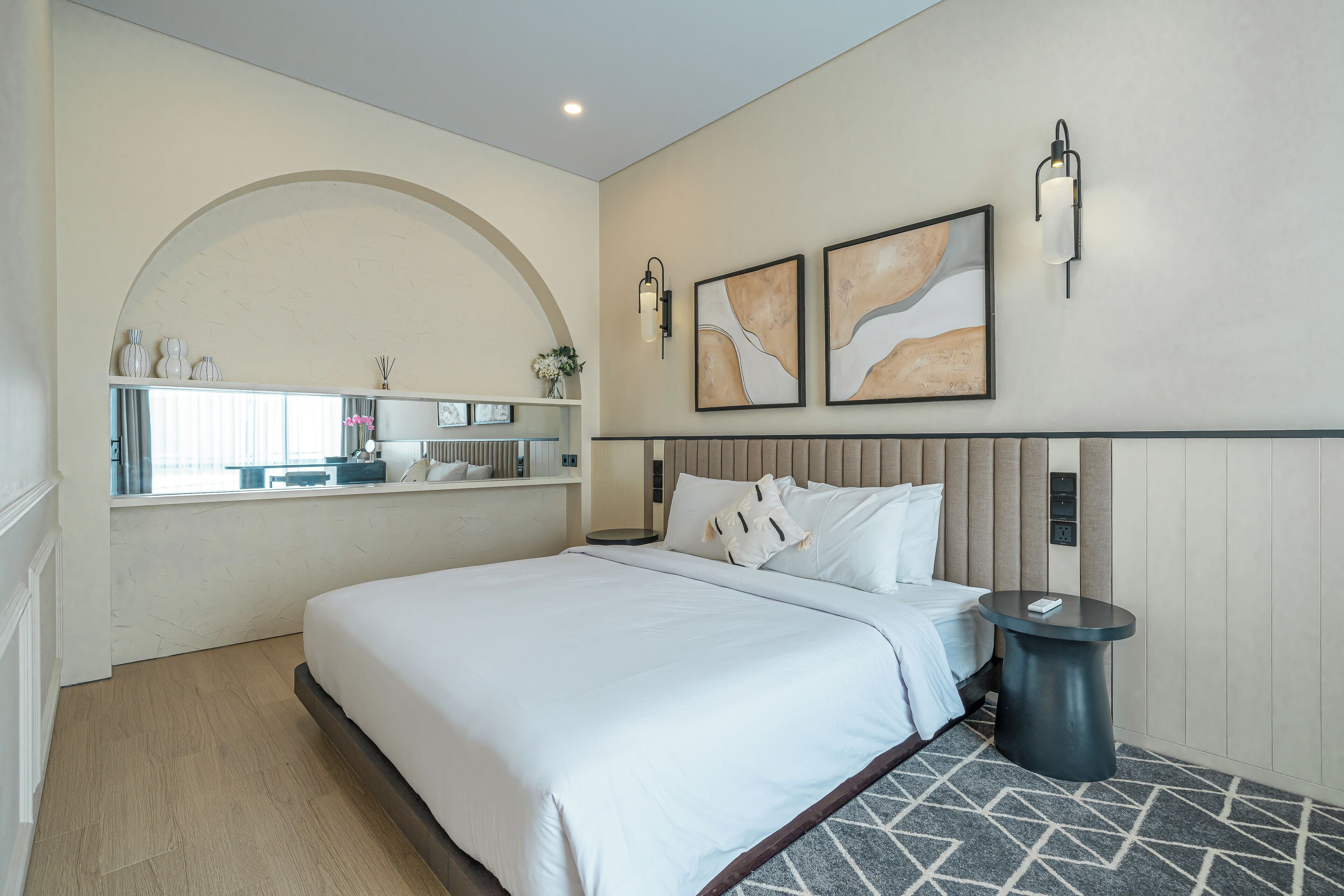 Una habitación de hotel moderna con una cama grande, una mesita redonda pequeña, un tocador con espejo y obras de arte en las paredes.