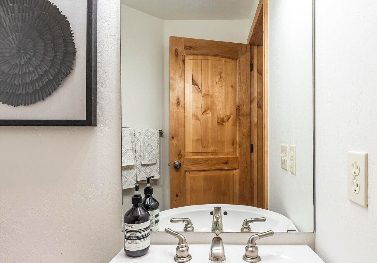 A bathroom with a wooden door, a white sink with two faucets, and a framed decorative piece on the wall.