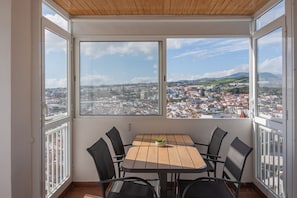 Un balcon avec une table et des chaises, de grandes fenêtres donnant sur la ville et un plafond en bois.