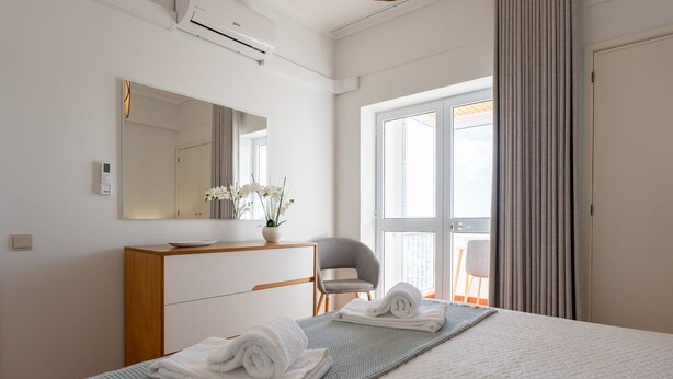 A hotel room with a bed, a desk, a chair, a mirror, and a window with curtains. - Solmar Sunset Place | Marina View (Ponta Delgada)