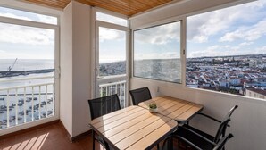A balcony with a wooden table and chairs, large windows with a city and port view. - Solmar Sunset Place | Marina View (Ponta Delgada)