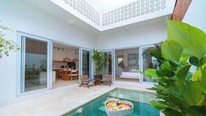 A modern indoor pool area with a dining setup and sliding glass doors leading to a kitchen. - Villa Arlo Canggu (Cemagi)