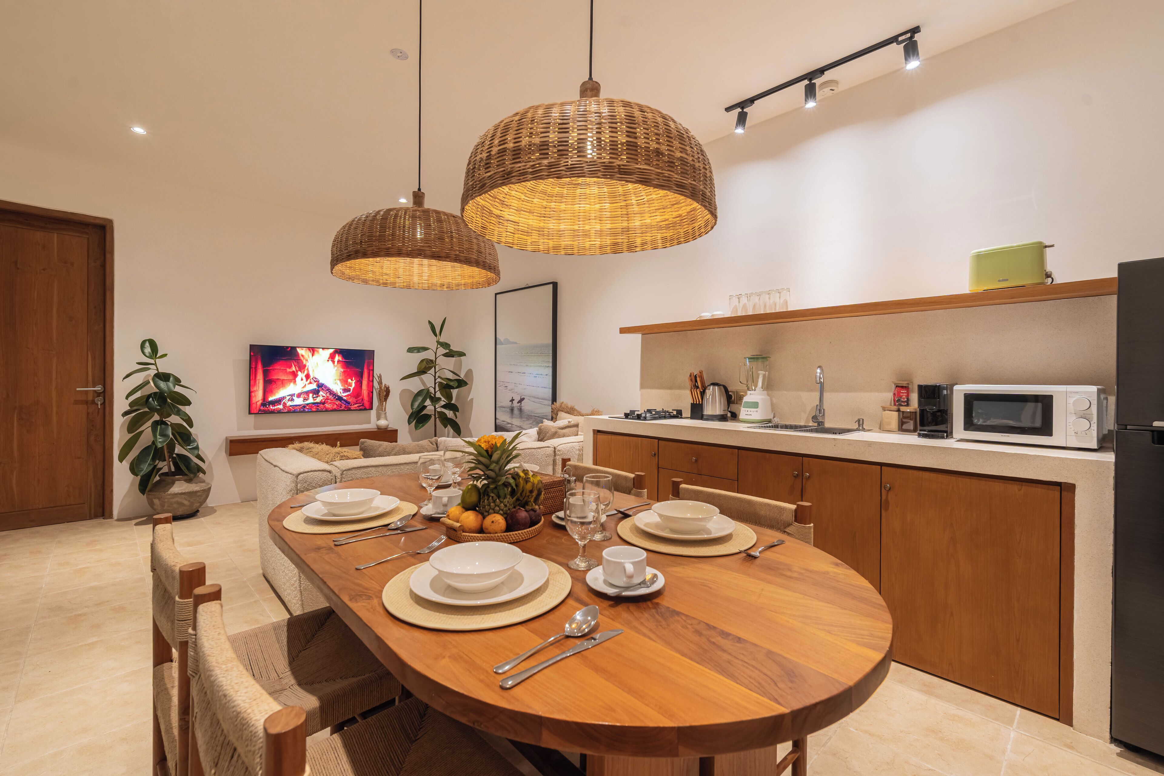 Ett modernt matområde med ett träbord för fyra personer, omgivet av flätade hänglampor, en tv som visar en brasa och ett kök med mikrovågsugn och kaffebryggare.