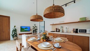 A dining area with a wooden table set for four, surrounded by wicker chairs. There are hanging wicker lamps above the table, a television mounted on the wall, and a kitchen area with a microwave and toaster visible. - Villa Arlo Canggu (Cemagi)