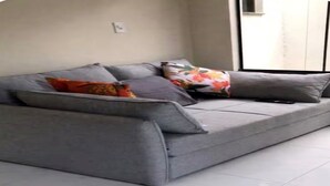 A grey sofa with multiple cushions in a room with white tiled flooring and a window.
