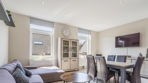Uma sala de estar moderna com um sofá, uma mesa de centro e um espaço de refeições com cadeiras e uma mesa. Há uma TV fixada na parede e um armário com portas de vidro.