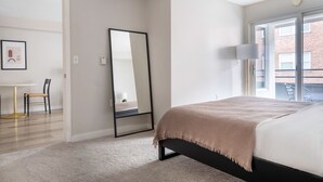 Ein Schlafzimmer mit einem Bett, einem großen Spiegel, einem Schreibtisch mit Stuhl und Blick auf ein Backsteinhaus durch ein Fenster.