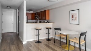 Eine moderne Küche mit Holzschränken, einer weißen Arbeitsplatte und einer eingebauten Mikrowelle. Drei schwarze Barhocker mit goldenen Füßen und ein runder Tisch mit zwei Stühlen stehen bereit.