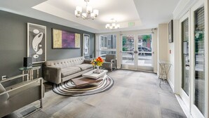 Eine moderne Lobby mit einem grauen Sofa, einem Couchtisch mit Blumen, einer Kristallleuchtre und einer Glastür zum Außenbereich.
