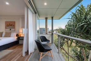 A bedroom with a bed, nightstands, and a painting. There is a balcony with chairs and a view of greenery. - Dune House (Tweed)