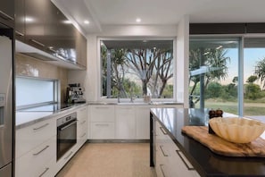 A modern kitchen with stainless steel appliances, a large window with a view of trees, and a wooden cutting board with a bowl on the countertop. - Dune House (Tweed)