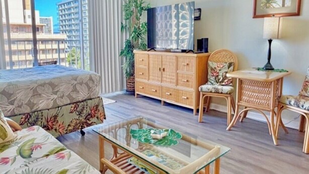 A hotel room with a bed, a glass-top table, a wooden cabinet, a wicker chair, and a view of buildings through a window. - Island-Inspired Cozy Retreat - Free WIFI (Honolulu)