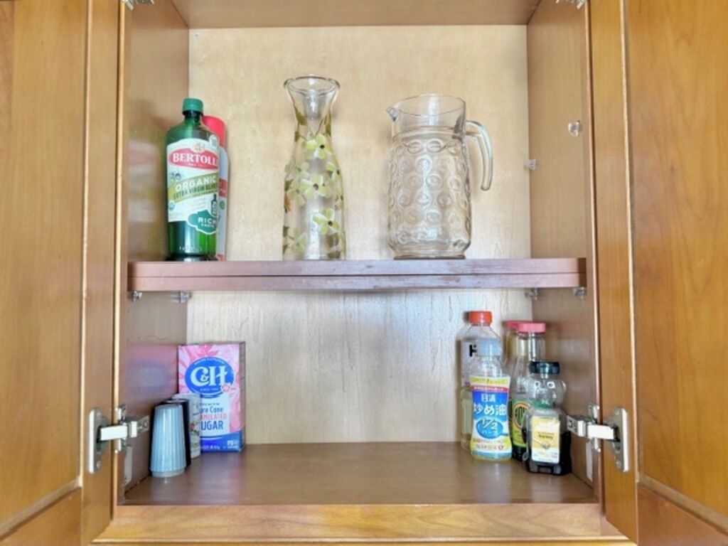 Ein Schrank mit einer Ablage, auf der eine Flasche Bertolli Bio-Essig, ein Glaskrug und eine Kanne stehen. Darunter befinden sich Flaschen mit C&H Zucker, ein Vorratsbehälter und eine Teedose.