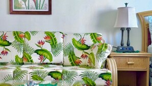 A room with a sofa featuring a tropical leaf pattern, a framed artwork on the wall, a bedside table with a lamp, and a wooden nightstand. - Island-Inspired Cozy Retreat - Free WIFI (Honolulu)