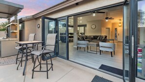 A modern house with a patio area featuring a table and chairs, a sliding glass door leading to a living room with a sofa and a ceiling fan. - Swim, Golf, Grill & Chill! Surprise Group Getaway (Surprise)