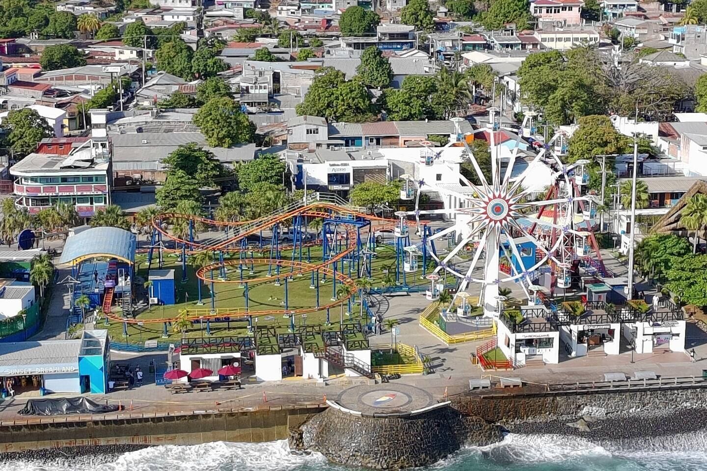 An aerial view of a coastal amusement park with a large Ferris wheel, roller coaster, and various rides.
