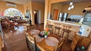 Un amplio espacio de sala con una mesa de comedor de madera, sillas y una zona de cocina con refrigerador y dispensador de agua.