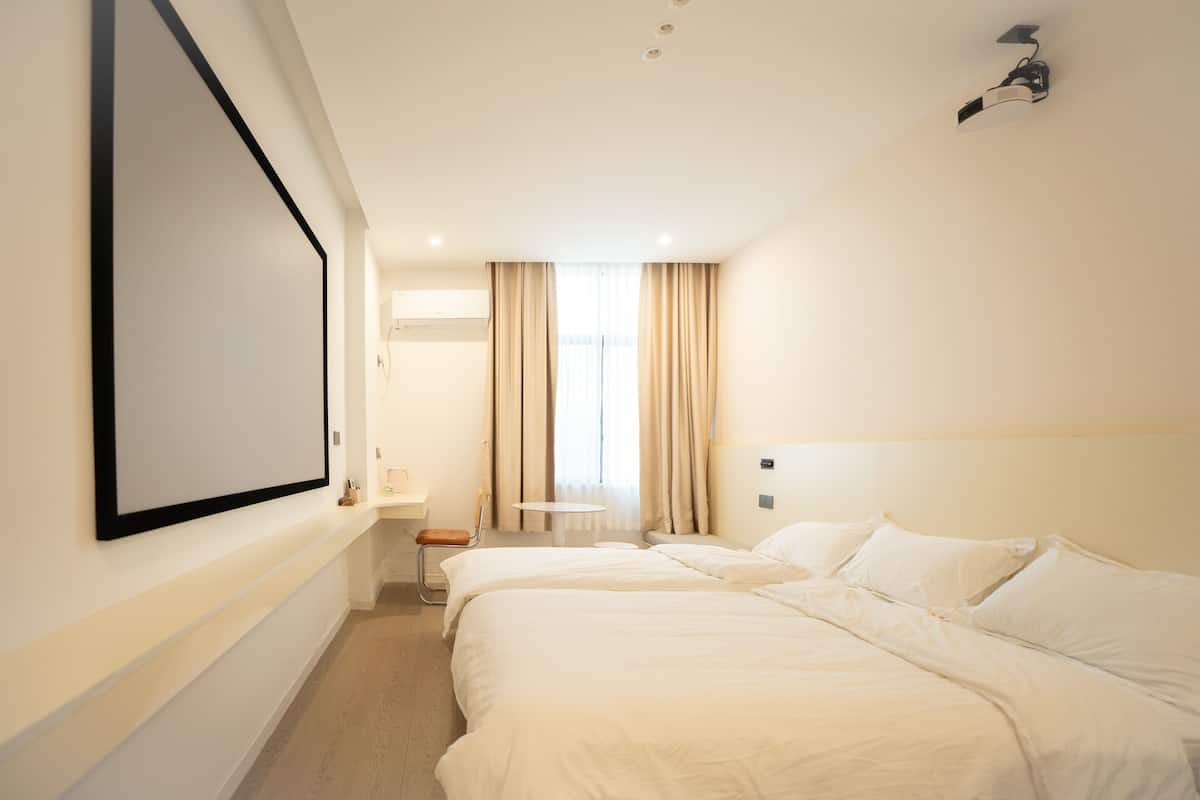 Habitación de hotel con dos camas, televisión montada en la pared, una mesita y una ventana con cortinas.