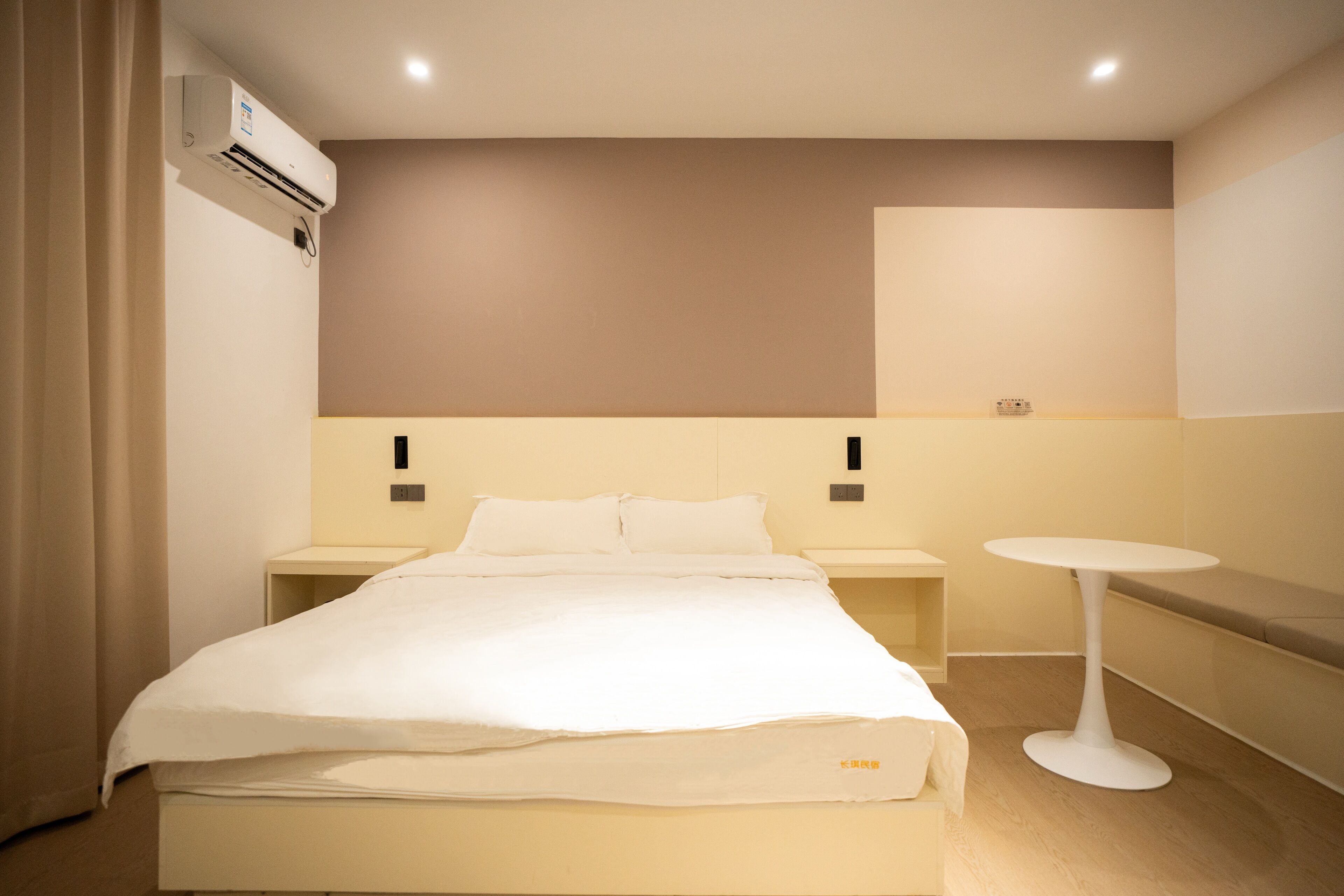 A hotel room with a bed, bedside tables, a round table, and an air conditioning unit.