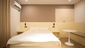 A hotel room with a bed, bedside tables, a round table, and an air conditioning unit.