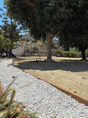 A gravel pathway leading to a shaded area with trees and a stone wall in the background. - Papyrise Oliveside (Heraklion)
