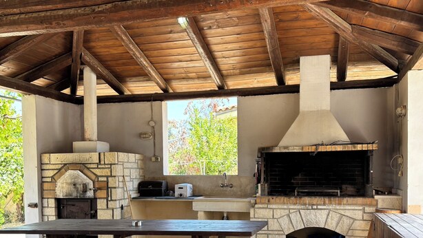 Uma cozinha rústica ao ar livre com forno de tijolos, churrasqueira e chaminé. Há mesas de piquenique e chão de concreto.