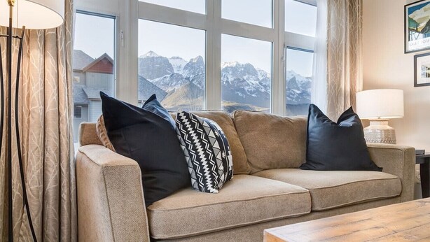 Ein gemütliches Wohnzimmer mit einem beige-farbenen Sofa, gemusterten Kissen und Blick auf schneebedeckte Berge durch große Fenster.