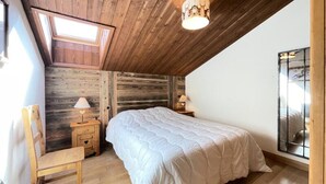 Un dormitorio con techo inclinado, paredes de madera, tragaluces, una cama con edredón blanco, dos mesitas de noche con lámparas y una ventana con celosía metálica.