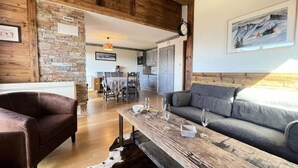 Una sala de estar acogedora con una pared de piedra, vigas de madera en el techo, un sofá, un sillón de cuero y un comedor al fondo.