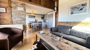 Un salón acogedor con una mesa de centro de madera, un sofá y un sillón de cuero. Se ve, en el fondo, una zona de comedor con una mesa y sillas.