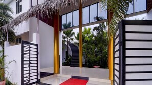 A modern building with a glass facade, a red carpet entrance, and a thatched roof structure. - Hotel O Auroville Bird Sanctuary Chennai (Vanur)
