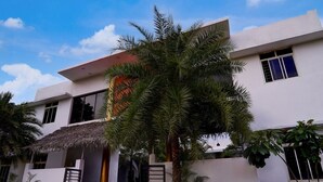 A modern two-story building with a flat roof, large windows, and a thatched roof entrance. - Hotel O Auroville Bird Sanctuary Chennai (Vanur)