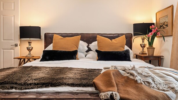 Ein ordentlich bezogenes Bett mit einer braun-weißen Bettdecke, zwei Nachttische mit Lampen, ein gerahmtes Bild an der Wand und ein Blumentopf auf dem Nachttisch.