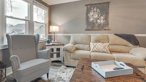 Een woonkamer met een zwart-wit ruit fauteuil, een gestreepte bank, een houten salontafel en een bloemrijk kunstwerk in een lijst aan de muur.