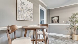 Een eethoek met een ronde tafel en twee houten stoelen, een groot bloemen schilderij aan de muur en een potplant.
