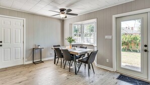 Un coin repas avec une table et des chaises en bois, un ventilateur de plafond et une vue sur l’extérieur, visible à travers une porte.