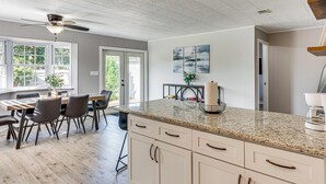 Une cuisine moderne dotée d’un plan de travail en granit, de meubles blancs et d’un ventilateur de plafond. On y trouve également un coin repas avec une table et des chaises en bois.