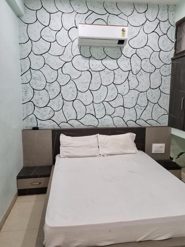 A bedroom with a patterned wall, a bed with white bedding, a nightstand, and a wall-mounted air conditioner. - Gokul Hotel (Malpura)