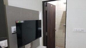 A room with a flat-screen TV mounted on a wall, a wooden door, and a tiled floor. - Gokul Hotel (Malpura)
