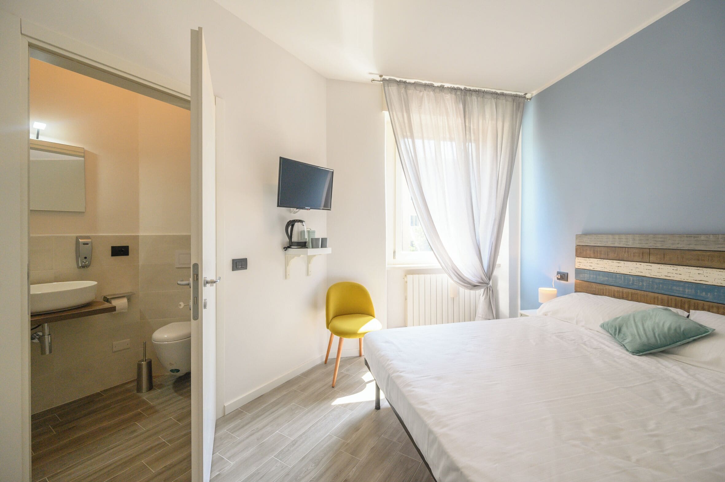 Quarto de hotel com cama, televisão, cadeira, banheiro com pia e janela com cortinas.