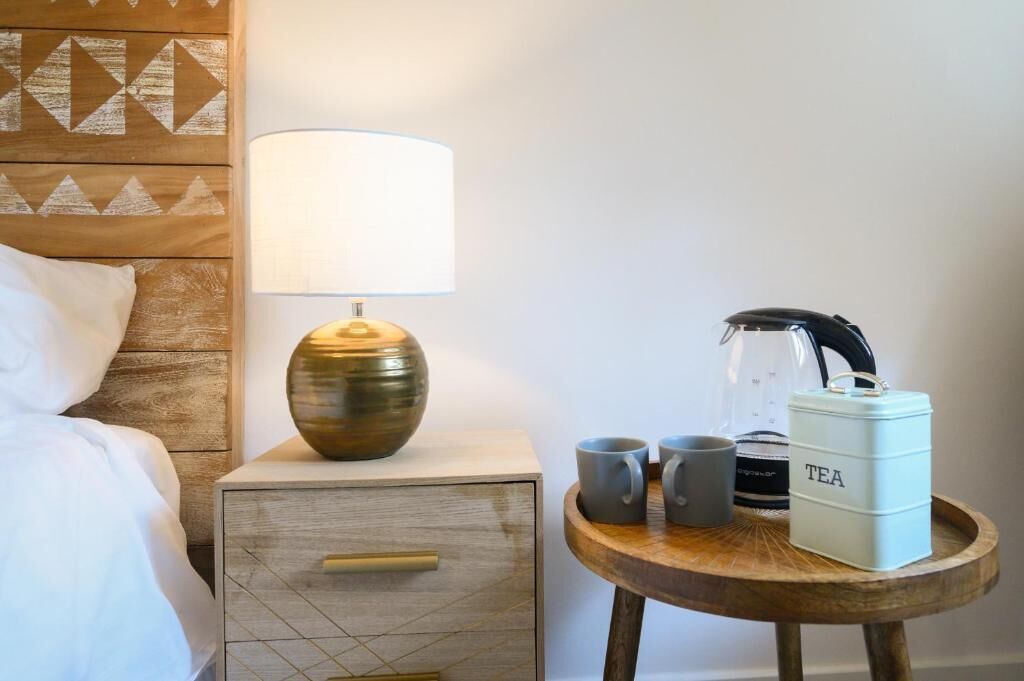 A neatly made bed with white linens, a bedside lamp, a small bedside table with a coffee maker, two mugs, and a tea container.