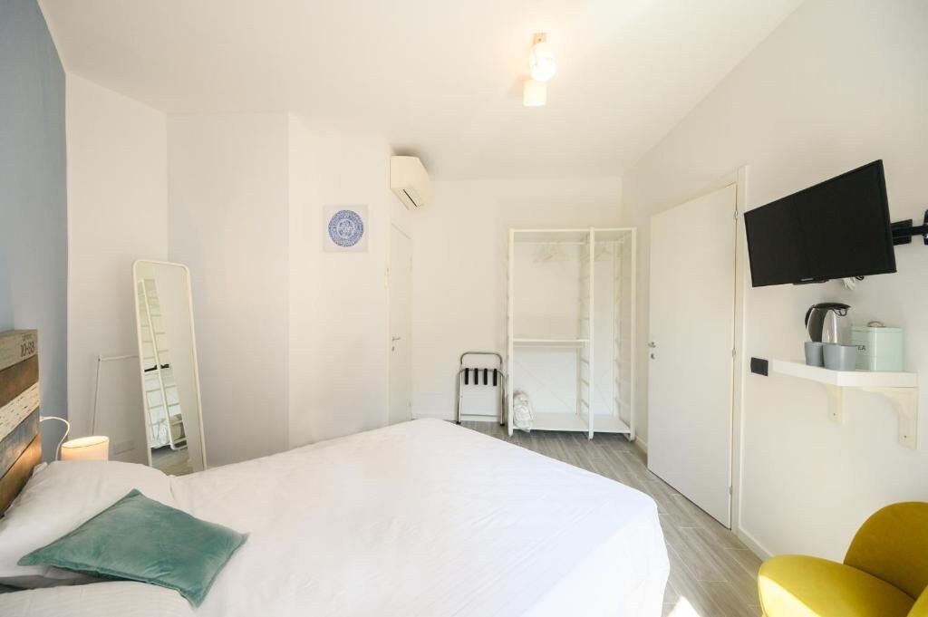 Habitación de hotel con una cama, televisión instalada en la pared, armario y un pequeño rincón de descanso con una silla.