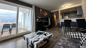 Ein modernes Wohnzimmer mit einer Couch, einem Couchtisch und einem Fernseher. Ein Balkon mit Stühlen bietet einen Blick ins Freie.
