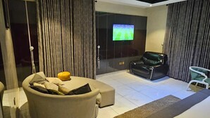 A modern living room with a beige sofa, a black leather chair, and a glass table. A flat-screen TV is mounted on the wall, and the room has a large window with curtains. - De Don's Villa (Abuja)