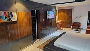 A modern bedroom with a large bed, a TV mounted on the wall, a wardrobe, and a seating area with a table and chairs. - De Don's Villa (Abuja)